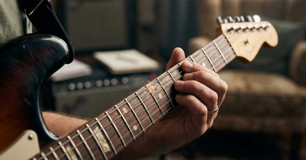 Close-up focado na escala de uma guitarra elétrica sunburst, mostrando uma corda vibrando e fazendo contato físico com um traste gasto, indicando trastejamento.