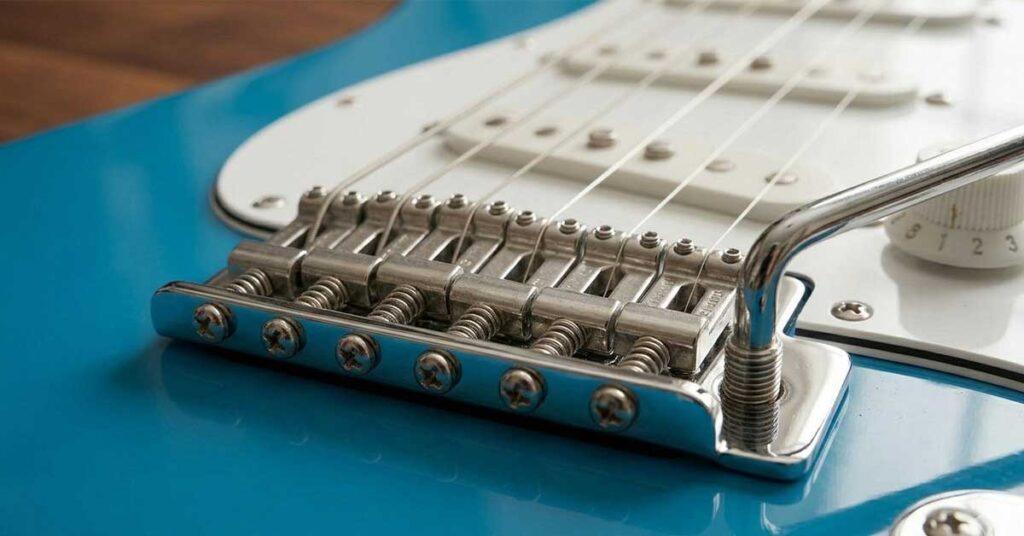 Close-up detalhado em ângulo de uma ponte móvel tremolo tradicional cromada com seis parafusos de montagem e alavanca (whammy bar) visível em uma guitarra azul brilhante.