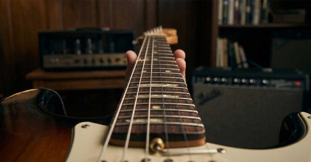 Visão em perspectiva de trás para a frente ao longo do braço de uma guitarra elétrica sunburst, focando no alinhamento das cordas e na curvatura da escala para regulagem do tensor.